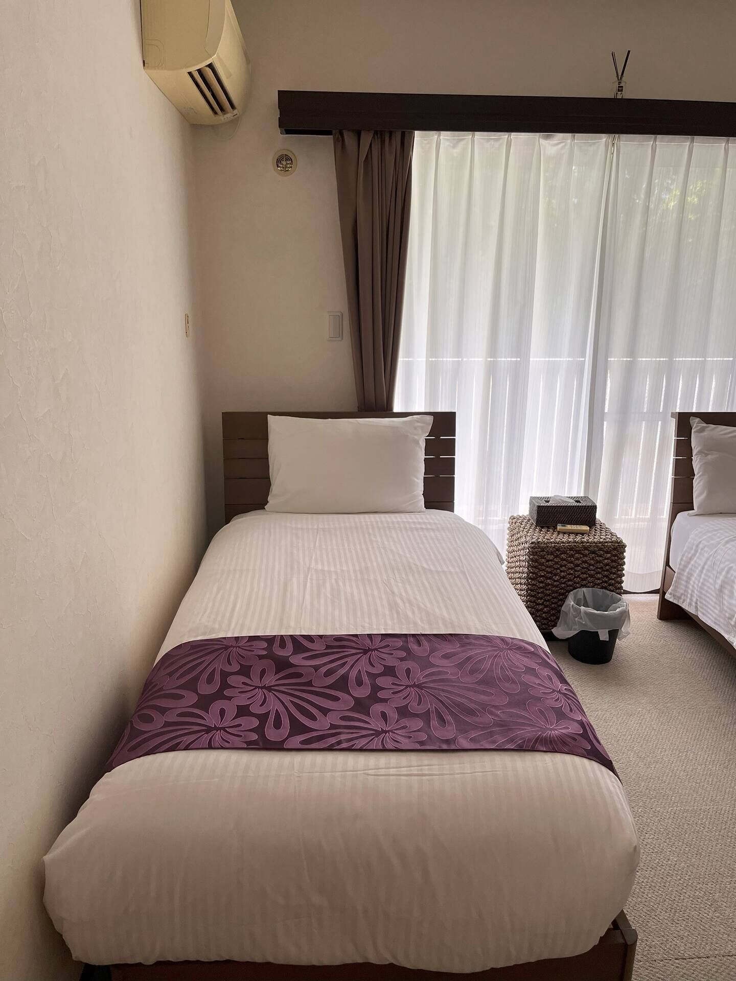A hotel room with a large bed, a smaller bed, a bedside table with a lamp, and a window with sheer curtains.