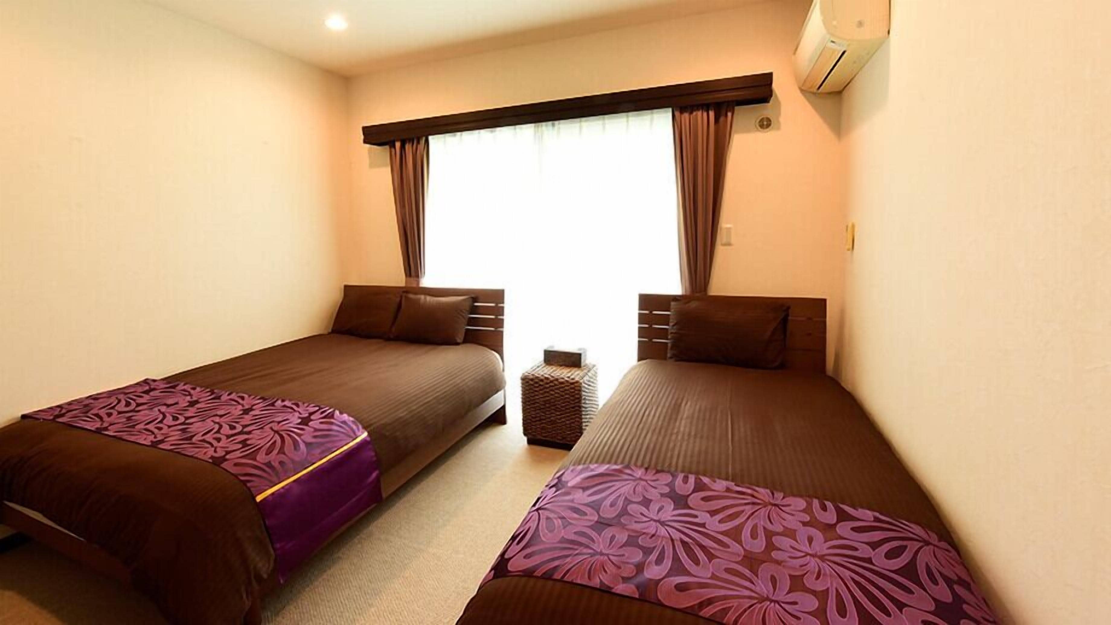 Una habitación de hotel con dos camas individuales, una mesita auxiliar y una ventana con cortinas.