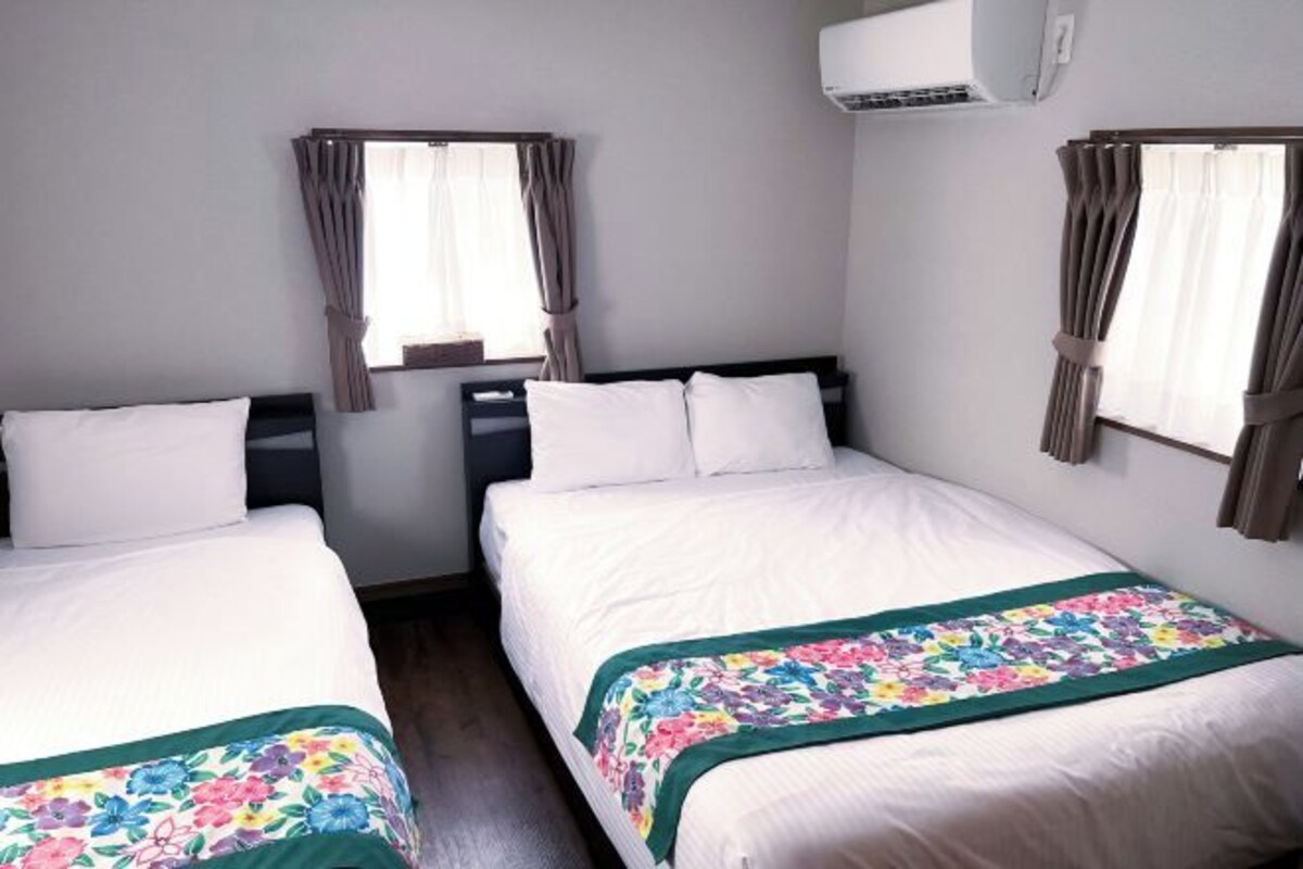 Habitación de hotel con dos camas individuales, una ventana con cortinas, un equipo de aire acondicionado y una colcha con motivos florales.