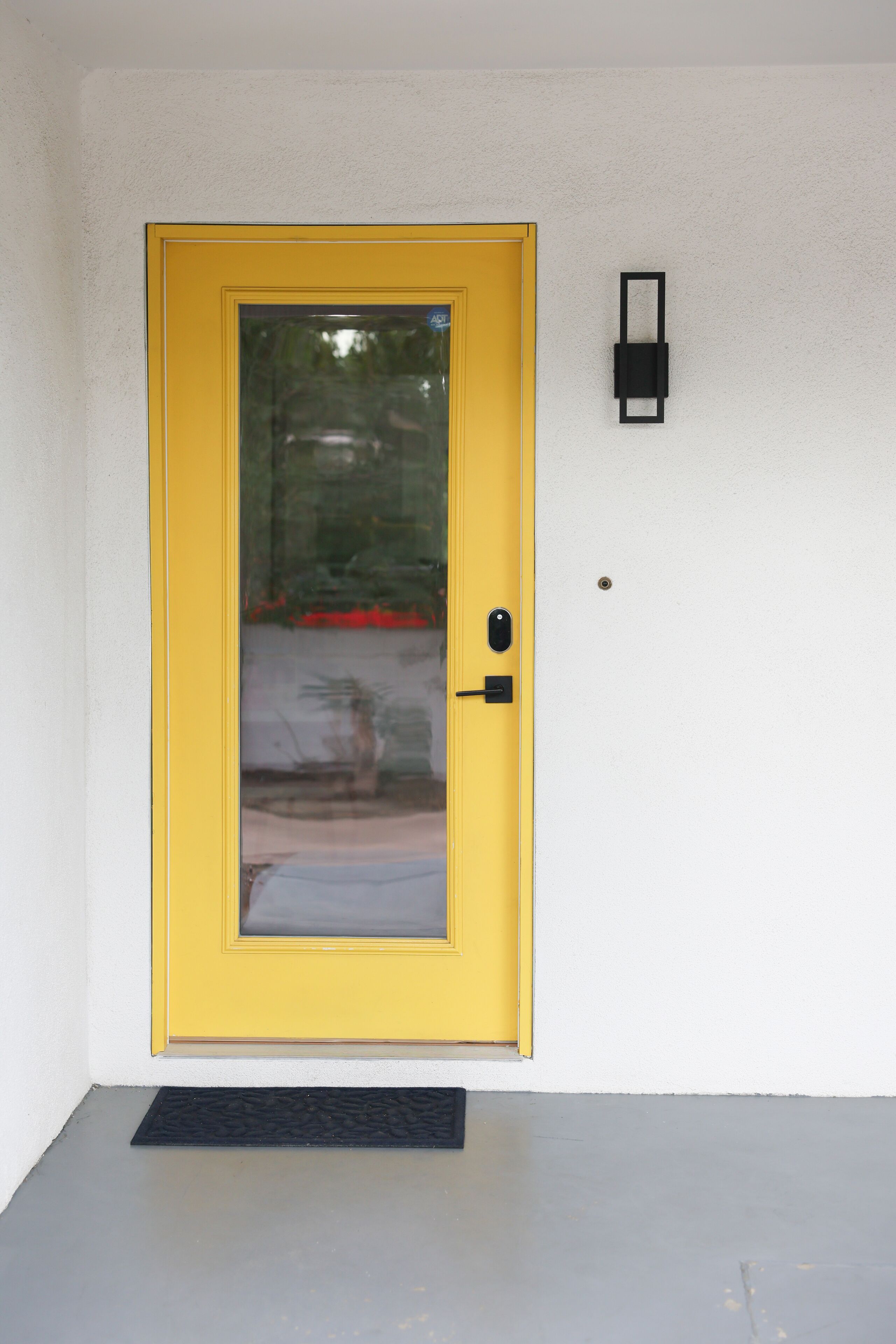 A yellow door with a glass panel, a black doorbell, and a black doormat on a concrete floor.