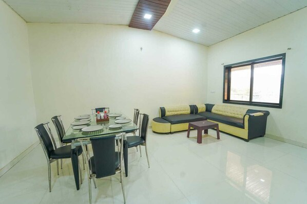 A spacious living area with a glass dining table, black chairs, a yellow and black sofa, and a small wooden table. - Lingmala Greens (Mahabaleshwar)