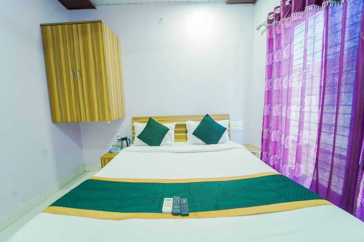 A single bed with a green and yellow bedspread, two green pillows, a wooden headboard, a wooden wardrobe, a nightstand with a kettle, and a purple curtain.