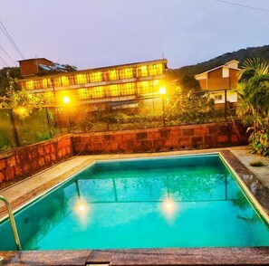 A rectangular swimming pool with a tiled edge, surrounded by a stone wall and a wooden deck. - Lingmala Greens (Mahabaleshwar)