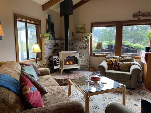 Una sala de estar acogedora con chimenea, dos sofás, una mesa de centro y ventanas amplias que ofrecen vista a la vegetación.