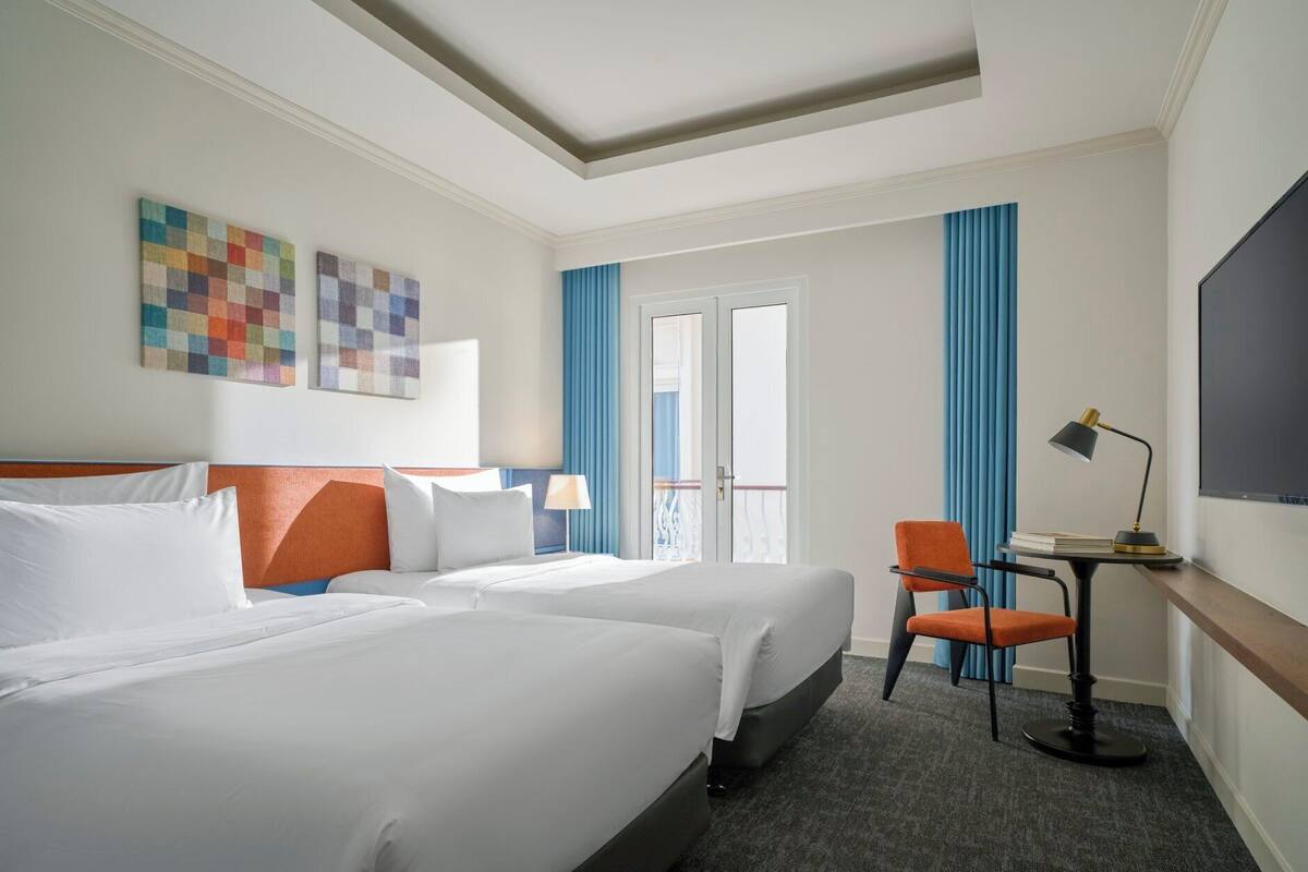 Une chambre d’hôtel avec deux lits, un bureau avec une chaise, une télévision et une fenêtre avec des rideaux.