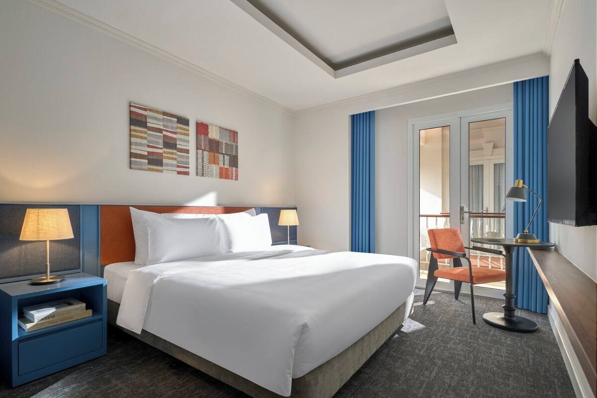 Une chambre d’hôtel avec un grand lit, deux tables de chevet avec des lampes, une chaise, une petite table avec une lampe, une télévision et un balcon avec des rideaux bleus.