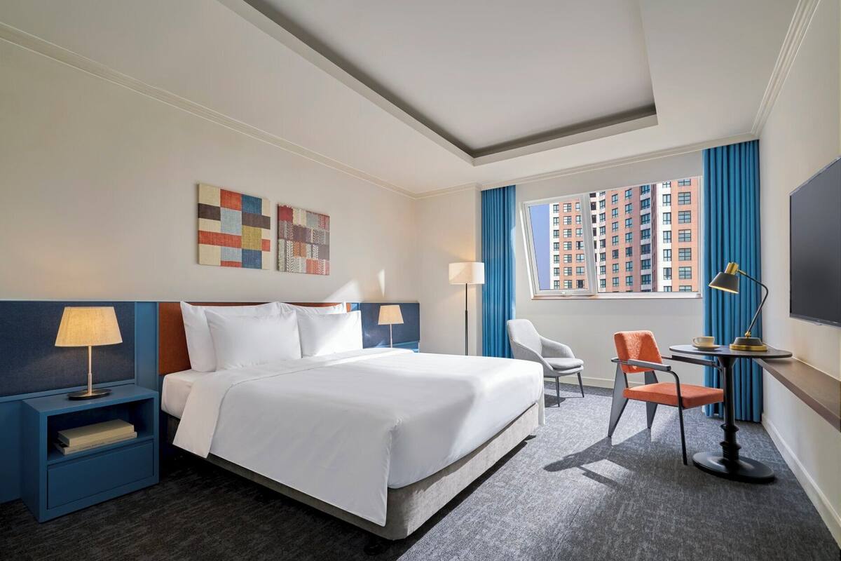 Une chambre d’hôtel avec un grand lit, deux tables de chevet avec des lampes, un bureau avec une chaise, une fenêtre avec des rideaux et des œuvres d’art accrochées au mur.