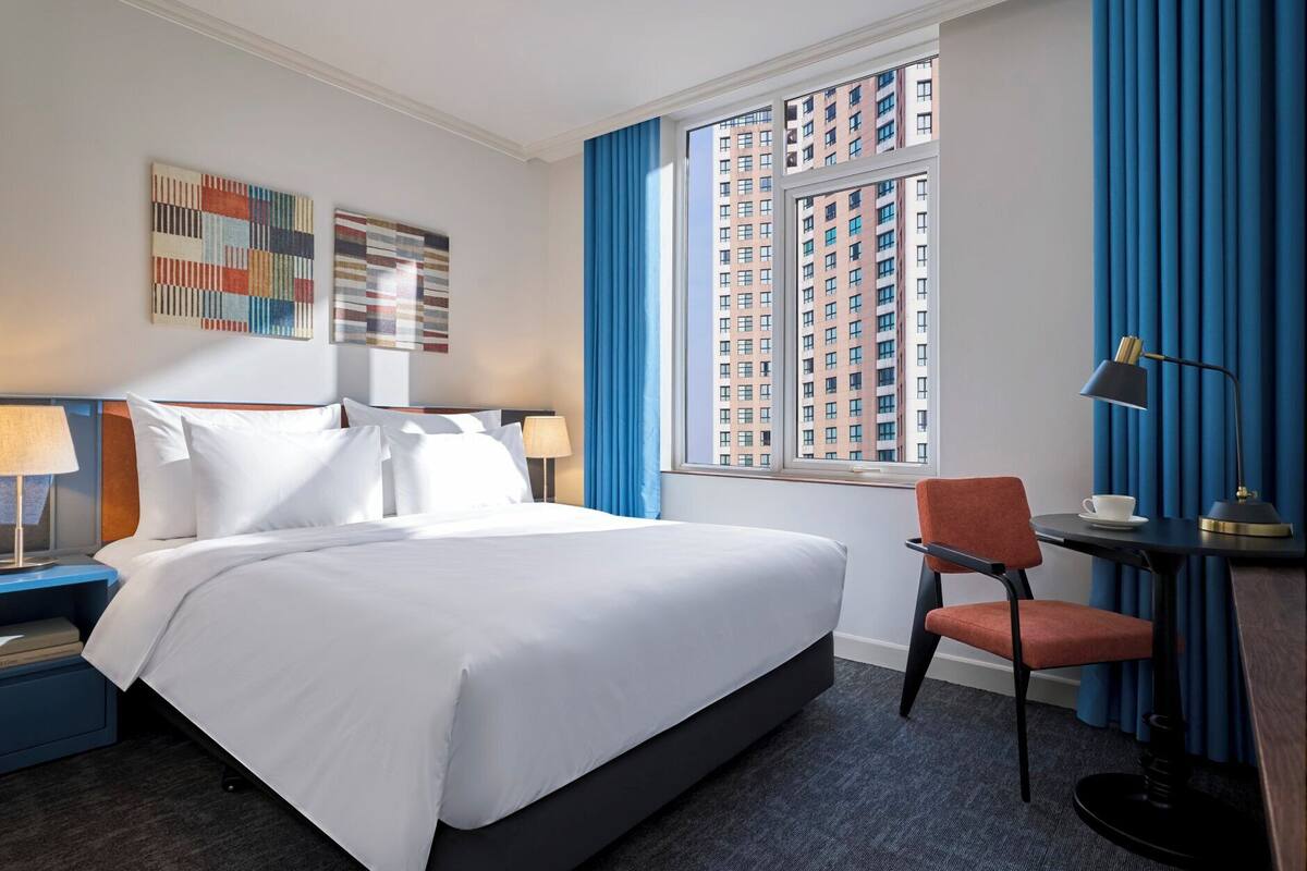 Une chambre d’hôtel avec un grand lit, un bureau avec une chaise, une fenêtre donnant sur des immeubles de grande hauteur et deux tableaux abstraits accrochés au mur.