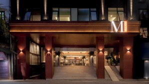 A modern hotel entrance with a prominent 'M' sign, a staircase leading to the lobby, and a well-lit facade.