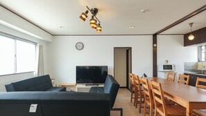 A modern living room with a black sofa, wooden dining table, and chairs. There is a flat-screen TV, a clock, and a kitchen area with a microwave. - Room 701 Gureisumukojima (Tokyo)