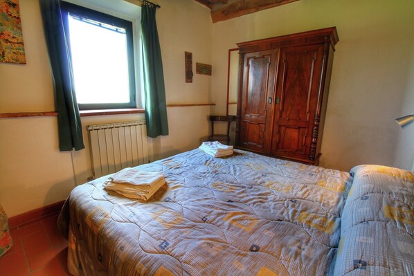 A bedroom with a bed, a wooden wardrobe, a radiator, and a window with curtains. - Farmhouse in Val d'Orcia Charm (Castiglione D'orcia)