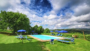 A swimming pool surrounded by blue lawn chairs and umbrellas, with a view of green hills and a partly cloudy sky. - Cottage in Tuscany with patio (Castiglione D'orcia)