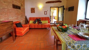 A cozy living room with a brick wall, a wooden bench, a sofa with colorful cushions, and a dining area with a table set for two. - Cottage in Tuscany with patio (Castiglione D'orcia)