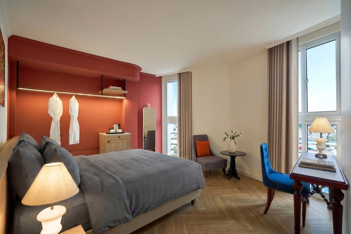 A hotel room with a bed, bedside table, lamp, chair, small table, and window.