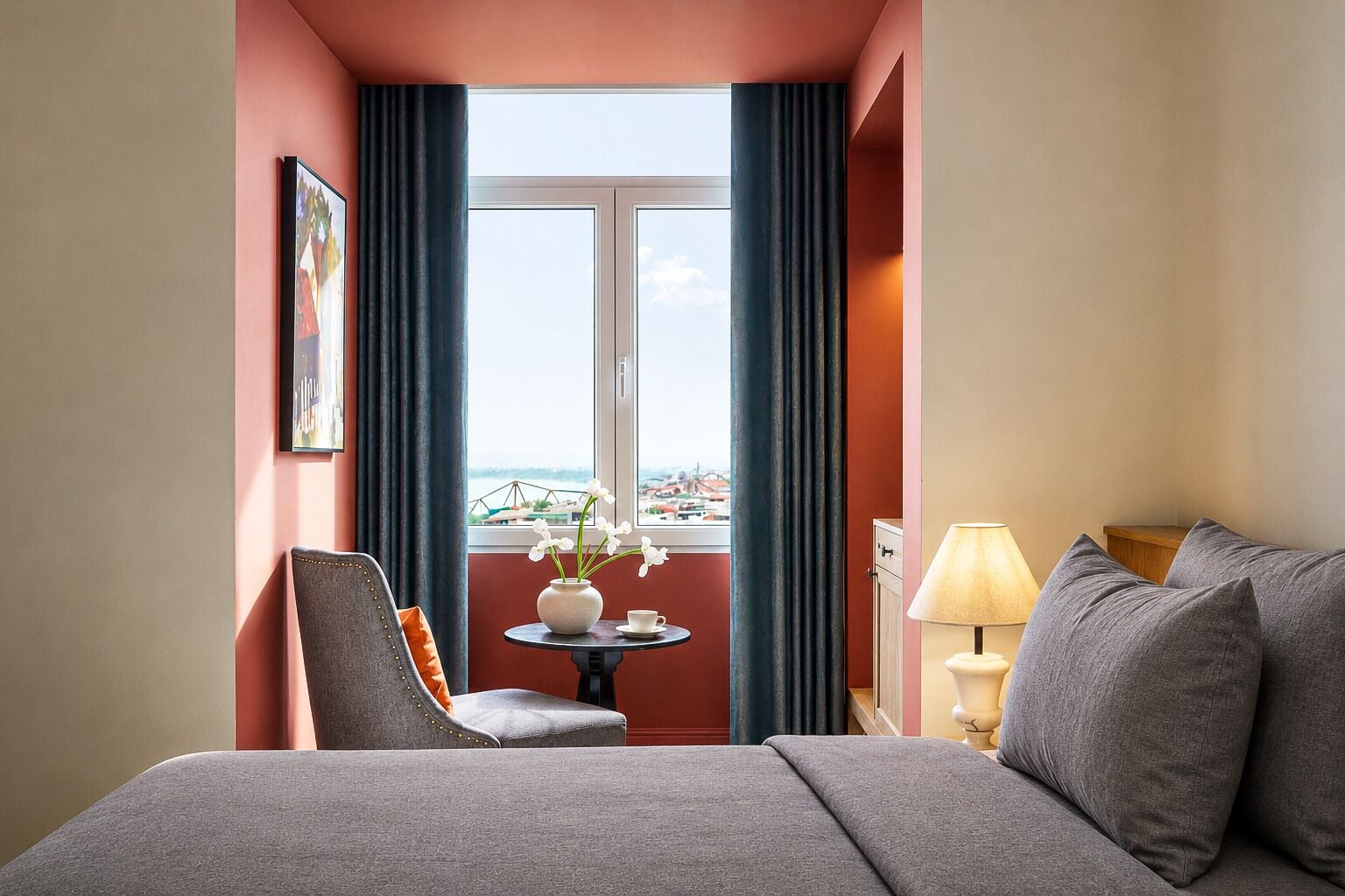 Une chambre d’hôtel avec un lit, une chaise, une table ornée d’un vase et de fleurs, une lampe, et une vue sur la ville par la fenêtre.