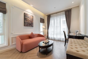 A living room with a sofa, a round coffee table, a chair, a bed, a painting on the wall, and a window with curtains. - M Village Hotel Long Biên, a brand of Modern Village Lifestyle (Hanoi)