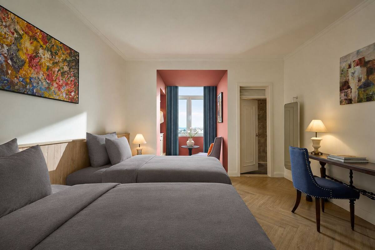 A hotel room with two beds, a desk, a chair, a window with curtains, and artwork on the walls.