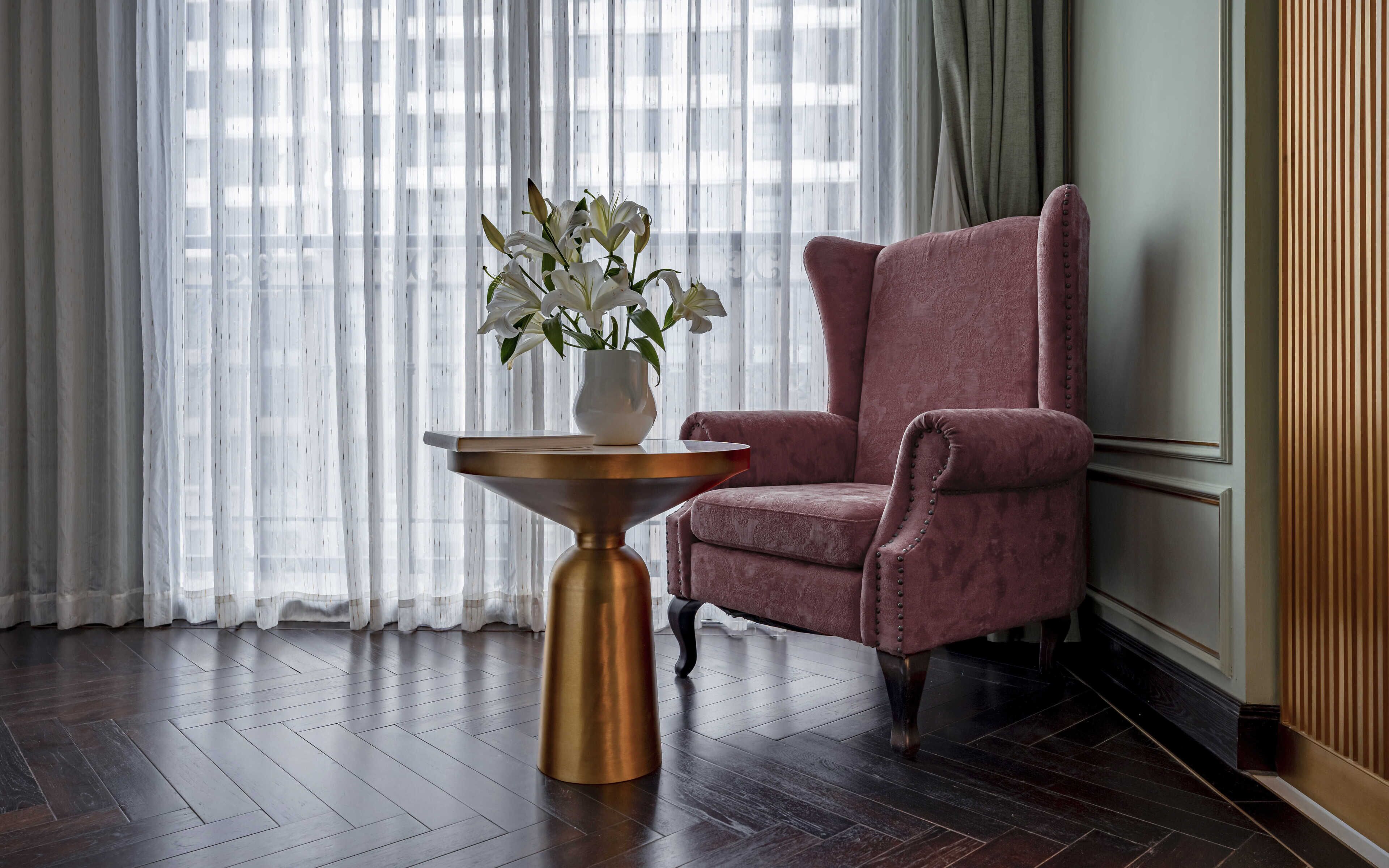 A room with a dark wood floor, a pink upholstered armchair, a small round table with a gold base, and a white vase with white flowers.