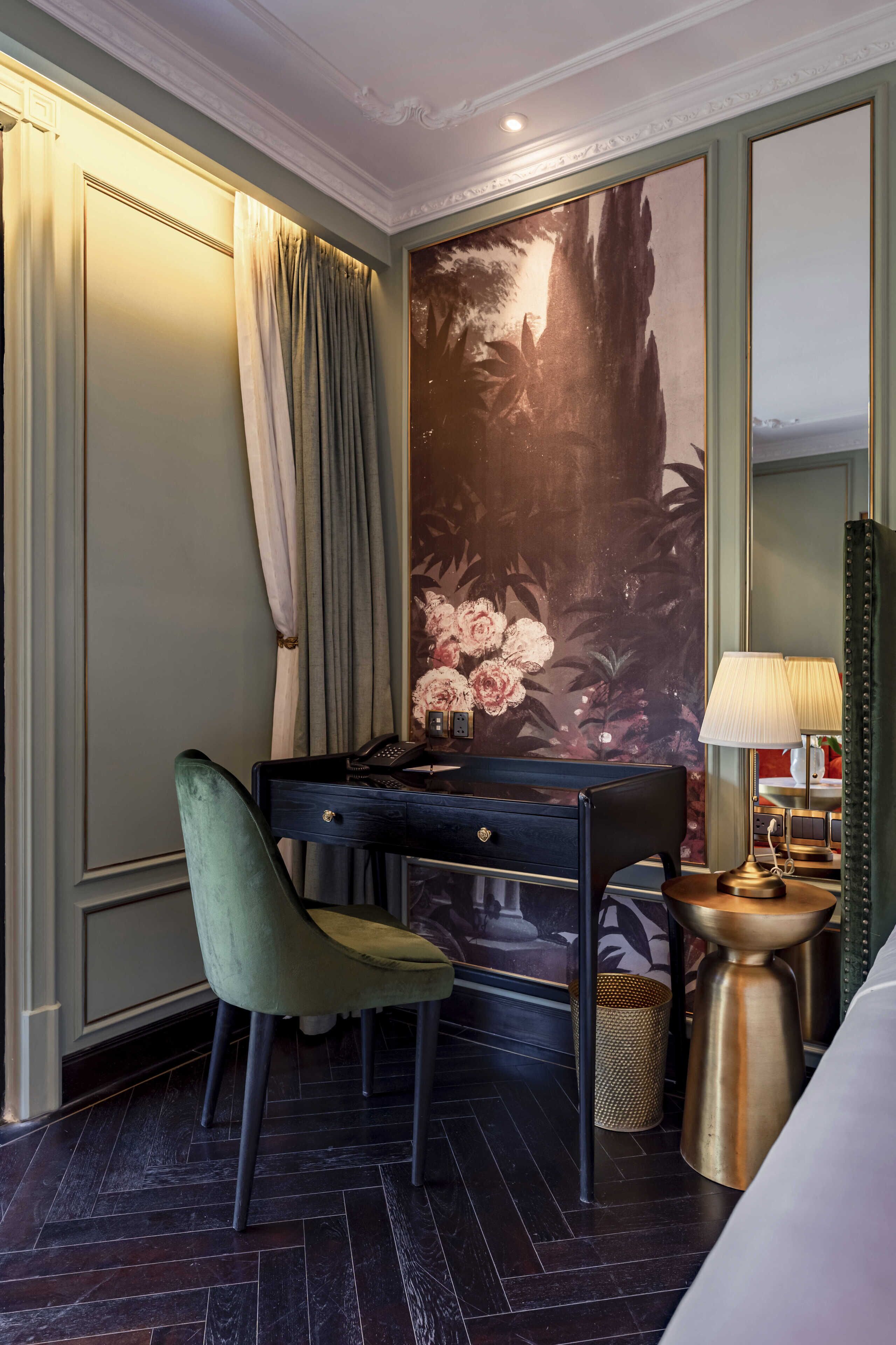 A hotel room with a desk, a green chair, a lamp, and a large floral mural on the wall.