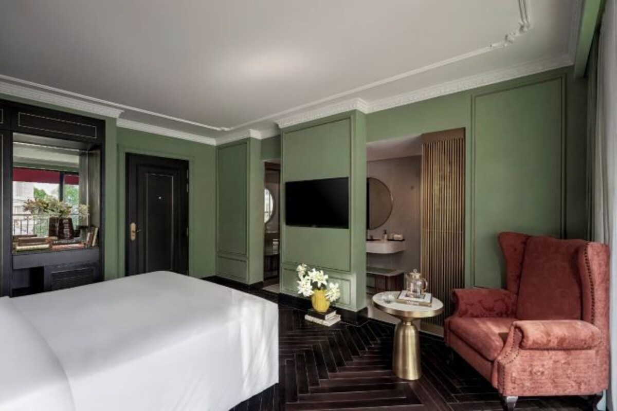 A hotel room with a large bed, a round table with a vase of flowers, a red armchair, and a television on the wall.