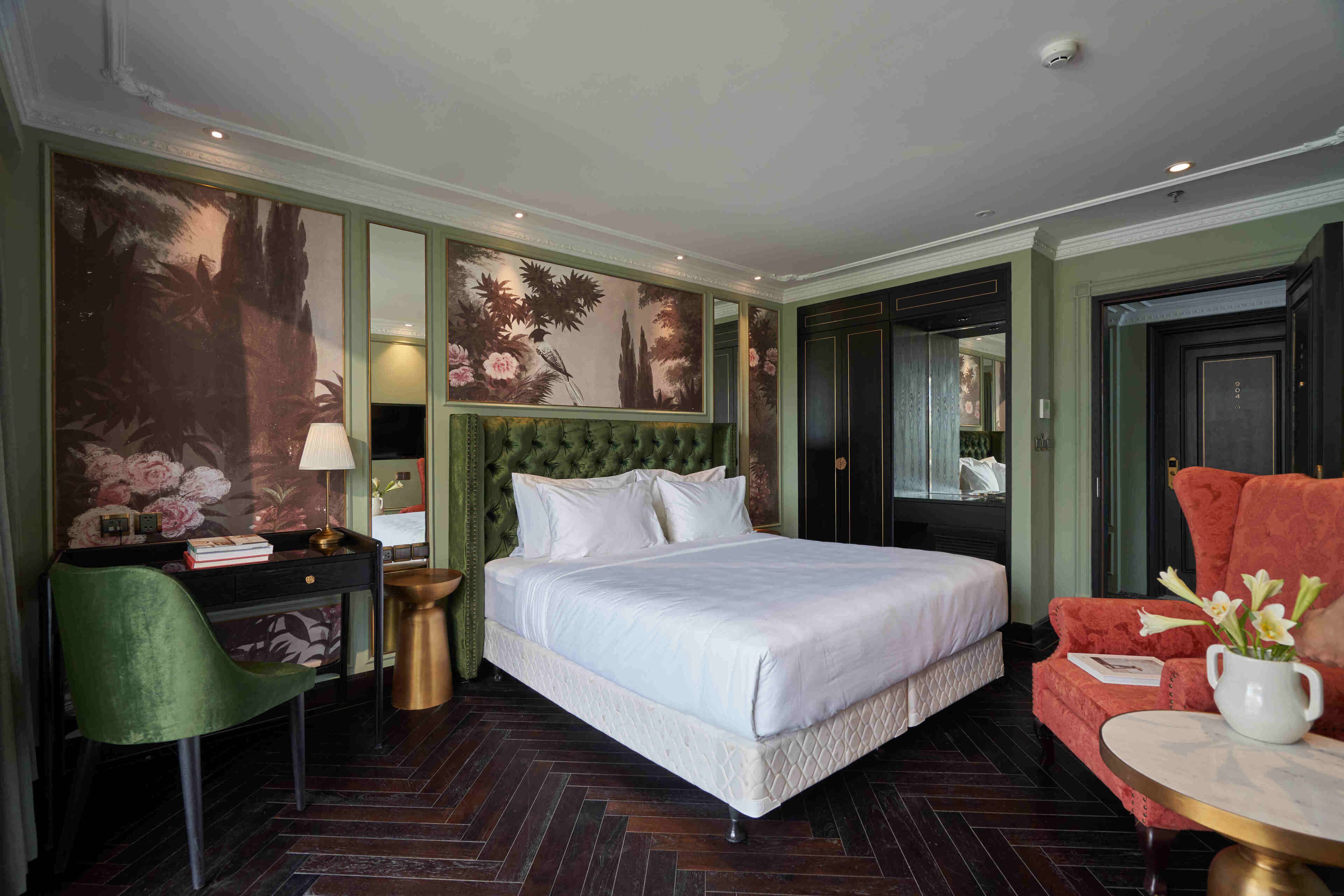 Une chambre d’hôtel avec un grand lit, un bureau avec une chaise, une table ronde avec un vase et un fauteuil rouge.