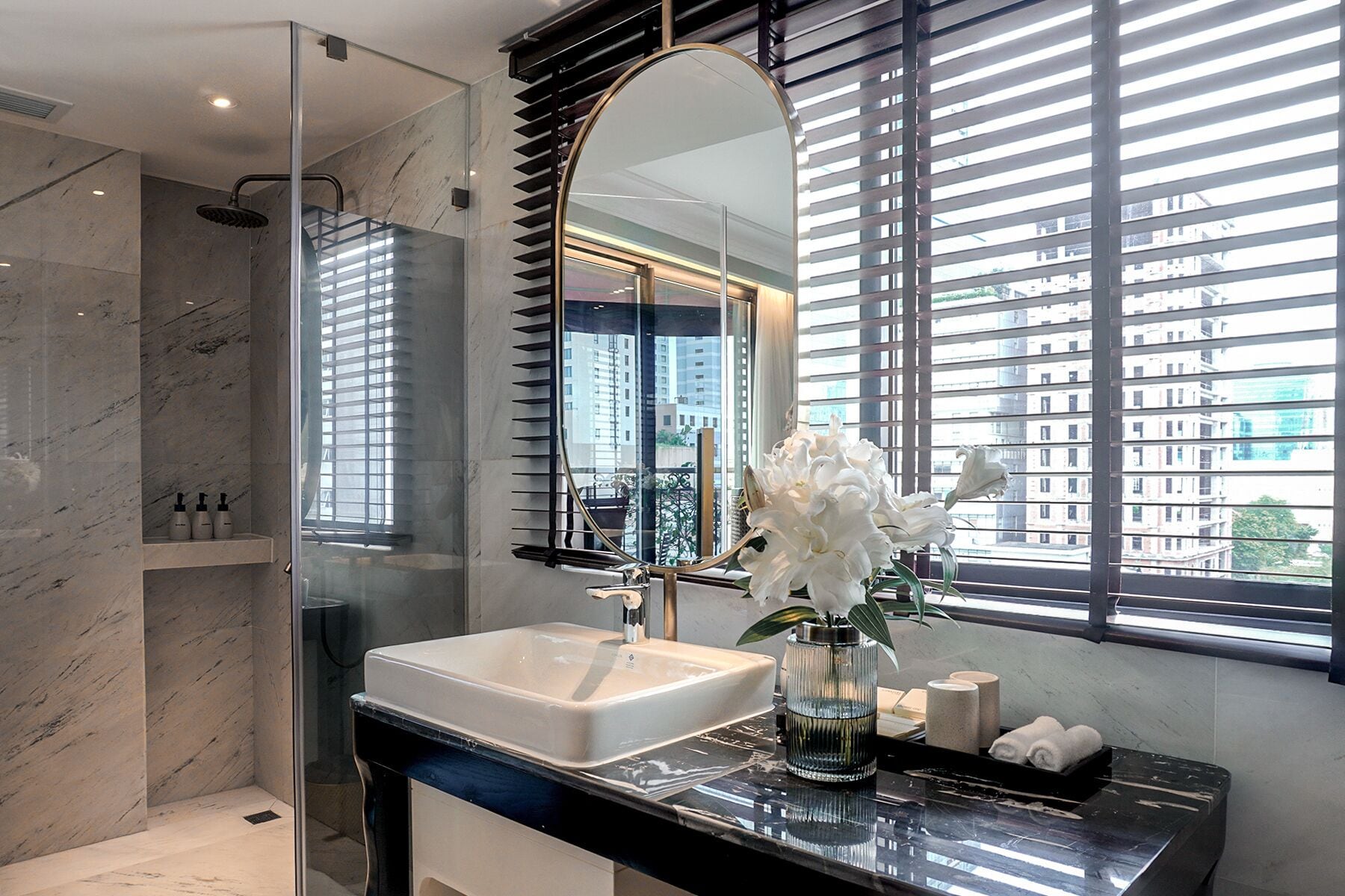 A modern bathroom with a marble sink, a large mirror, and a view of the city through floor-to-ceiling windows.