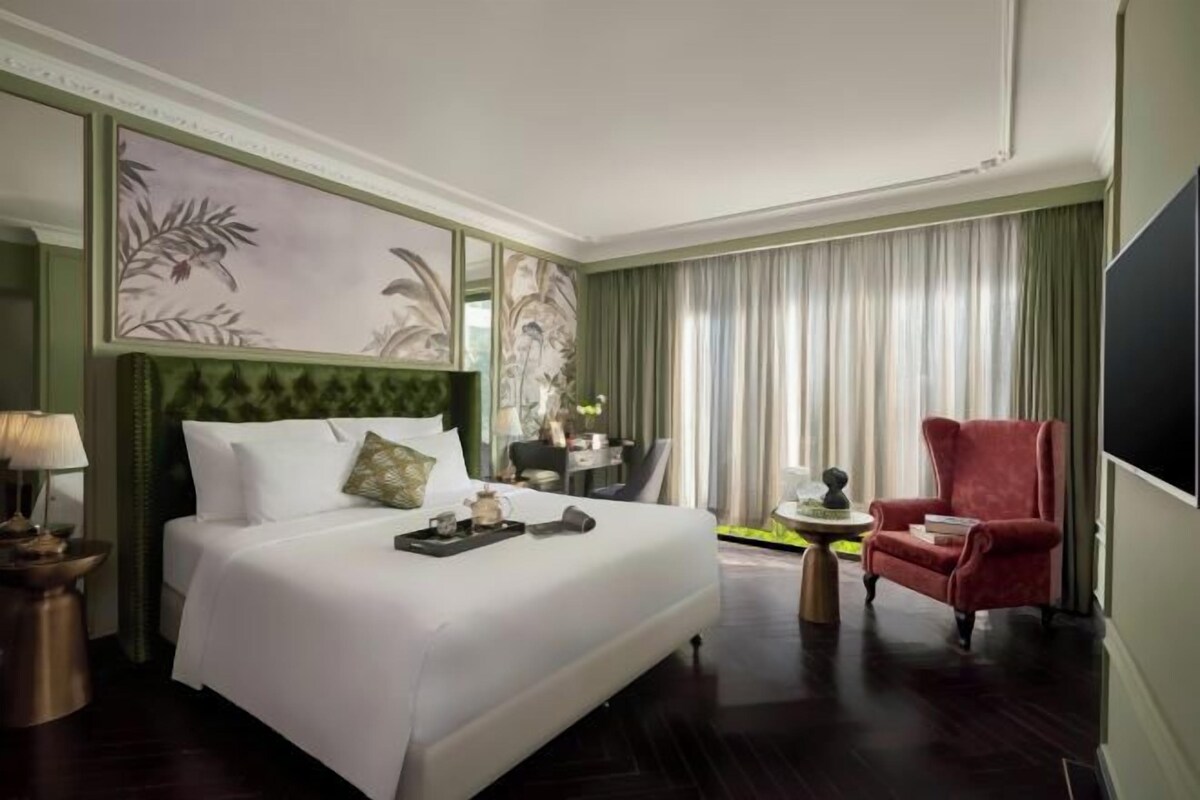 A hotel room with a large bed, a red armchair, a desk with a chair, and a large mirror on the wall.