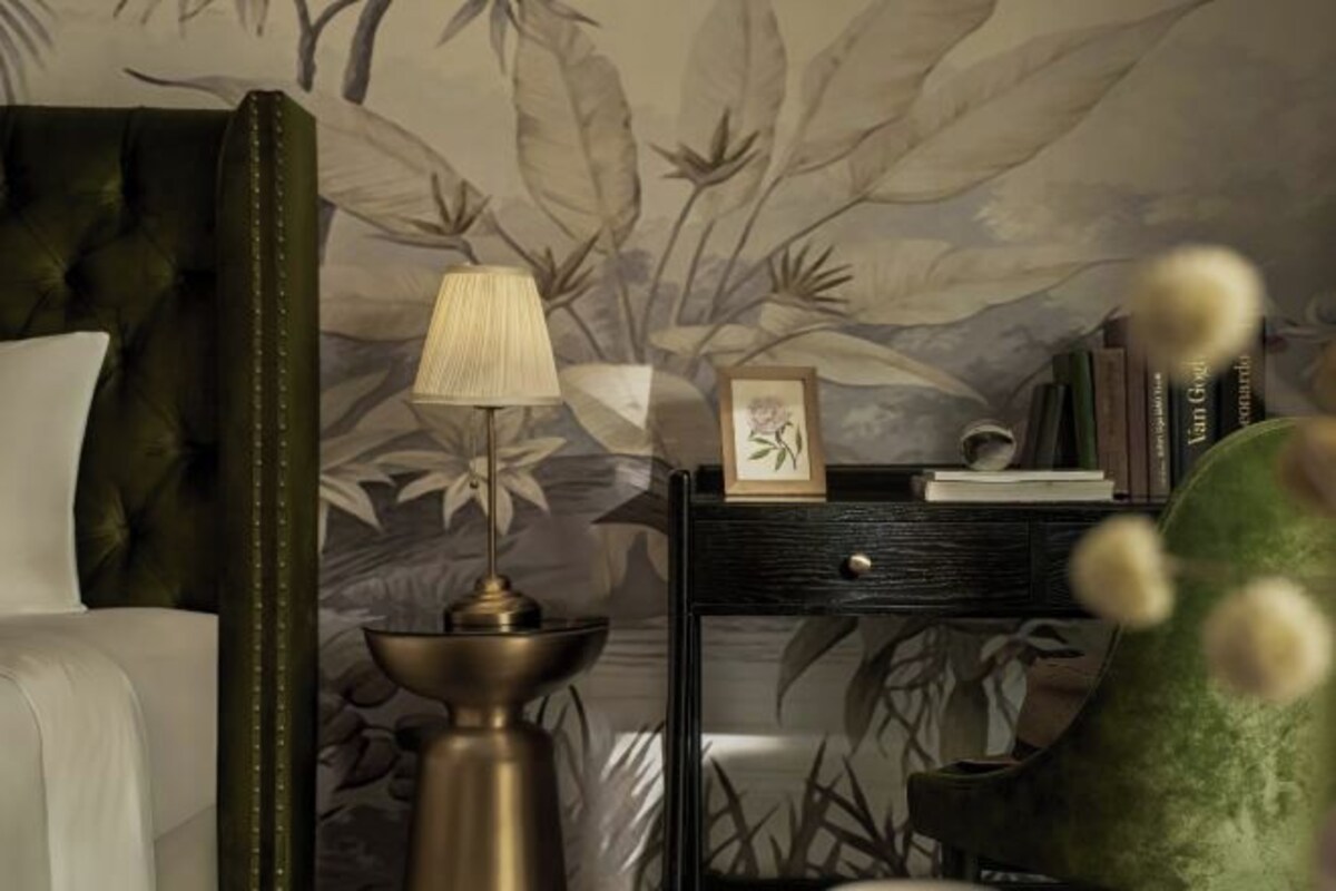 Un dormitorio con cabecera capitonada, una lámpara de noche, un cuadro enmarcado y una mesita de noche con libros y un florero.
