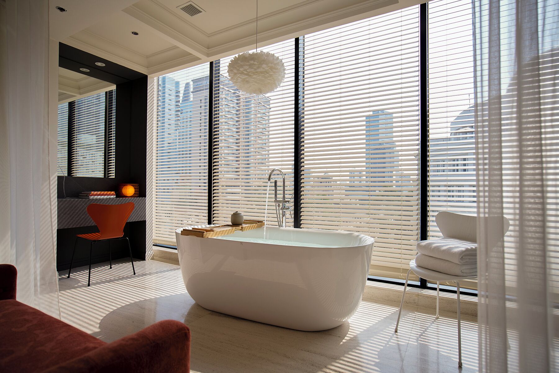A modern bathroom with a freestanding bathtub, a large window with blinds, and a view of the cityscape.