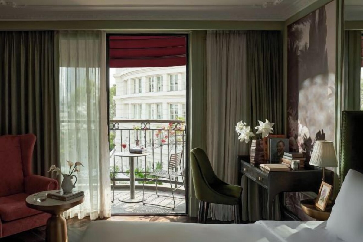 A hotel room with a bed, a desk with a lamp, a chair, a small table with a vase of flowers, and a view of a building through a balcony door.