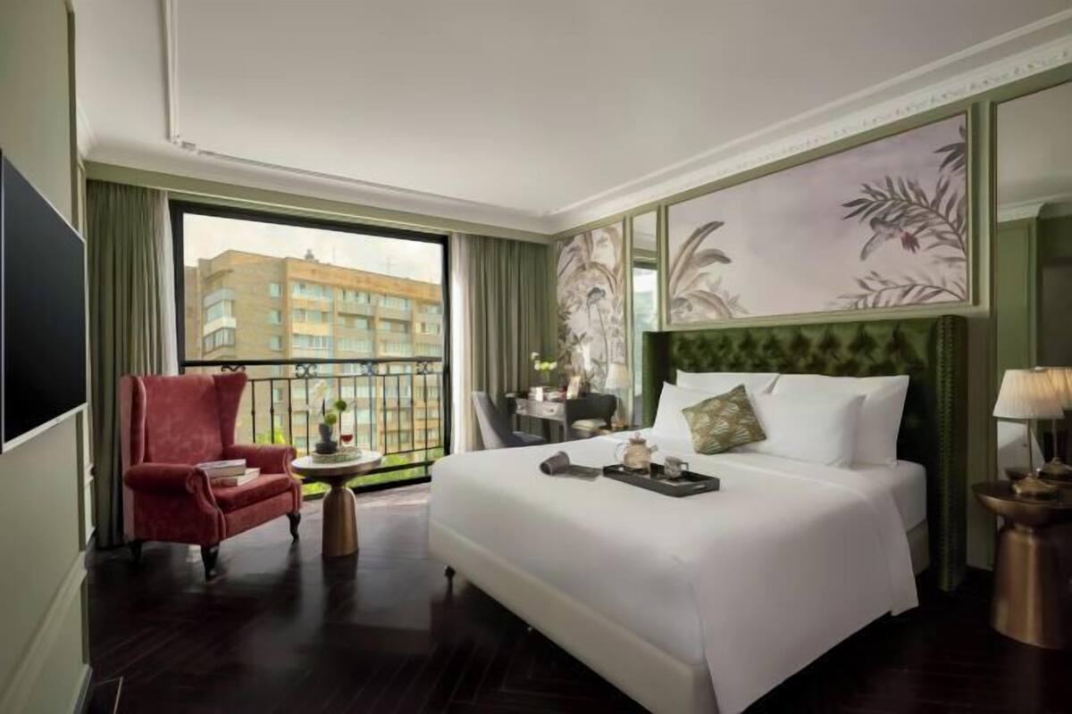 Habitación de hotel con una cama grande, un sillón rojo, una mesita con una bandeja y vista a un edificio desde la ventana.