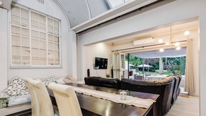 A dining area with a long table, chairs, and a sofa. There is a TV mounted on the wall and a window with blinds.