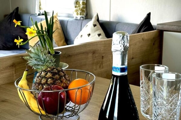 A dining table with a fruit basket containing bananas, pineapple, and an orange, a bottle of champagne, and two crystal glasses.