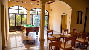 A billiards room with a pool table, wooden chairs, and large windows offering a view of greenery.