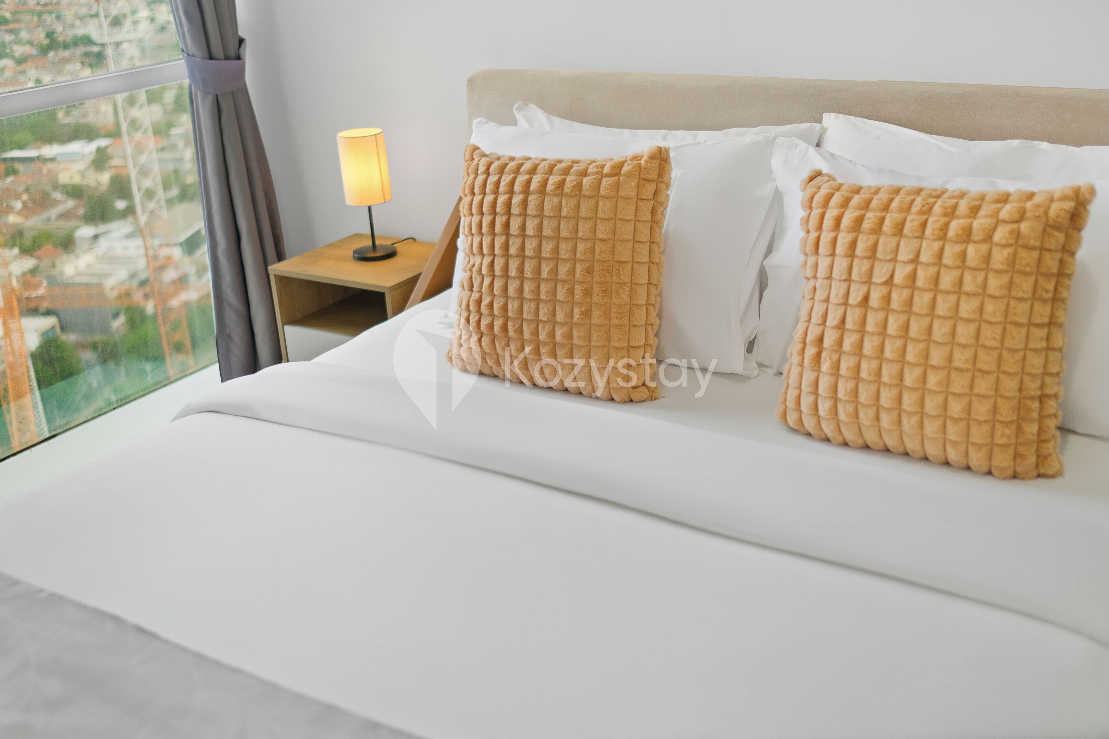 A hotel room with a bed, two textured pillows, a nightstand with a lamp, and a window with a view of buildings.