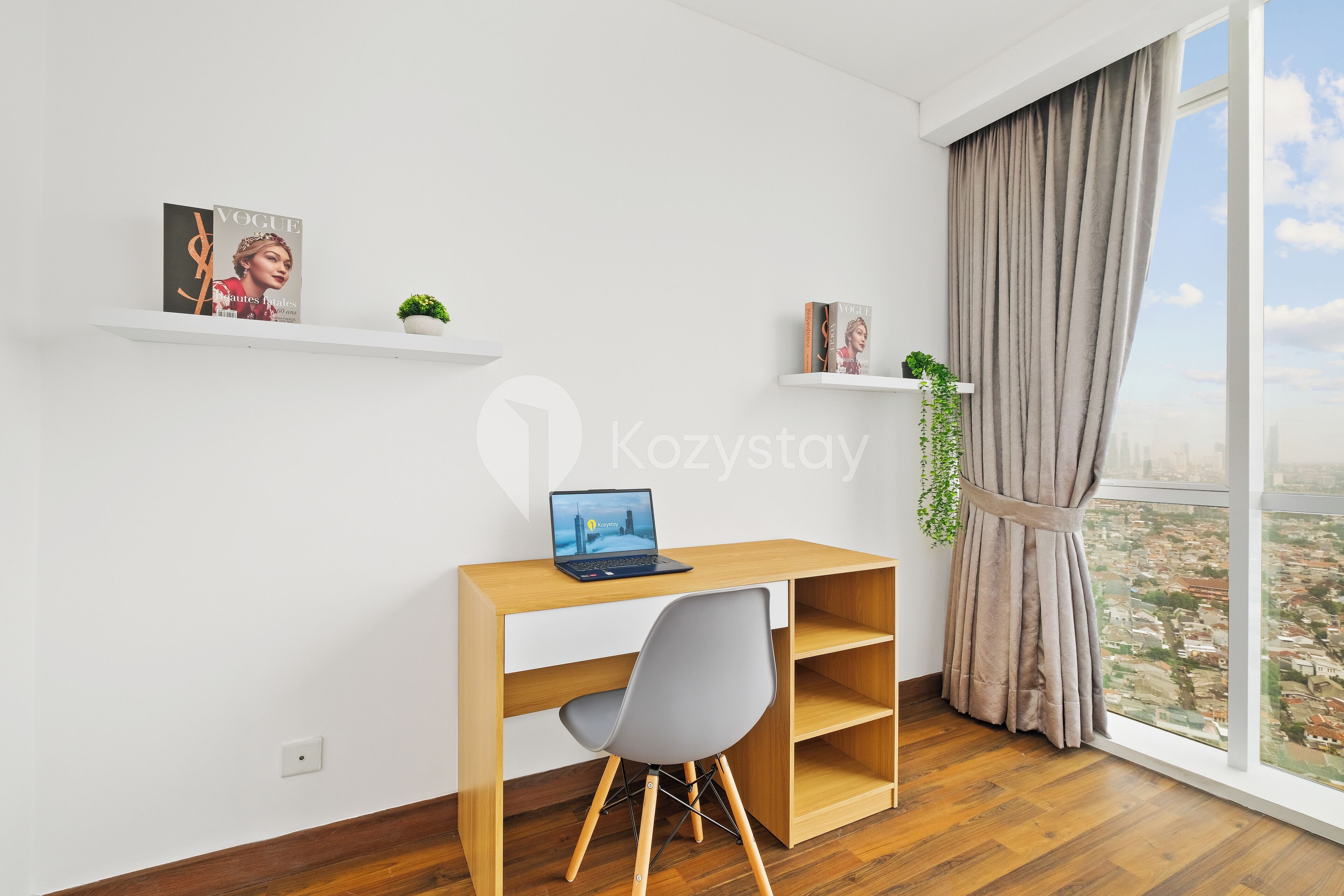 Une chambre moderne avec un bureau en bois, un ordinateur portable, une chaise grise et une vue sur la ville à travers de grandes fenêtres.