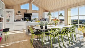 Een ruime keuken met houten plafond en grote ramen, een eethoek met groene stoelen en een ronde tafel, en uitzicht op de omgeving.