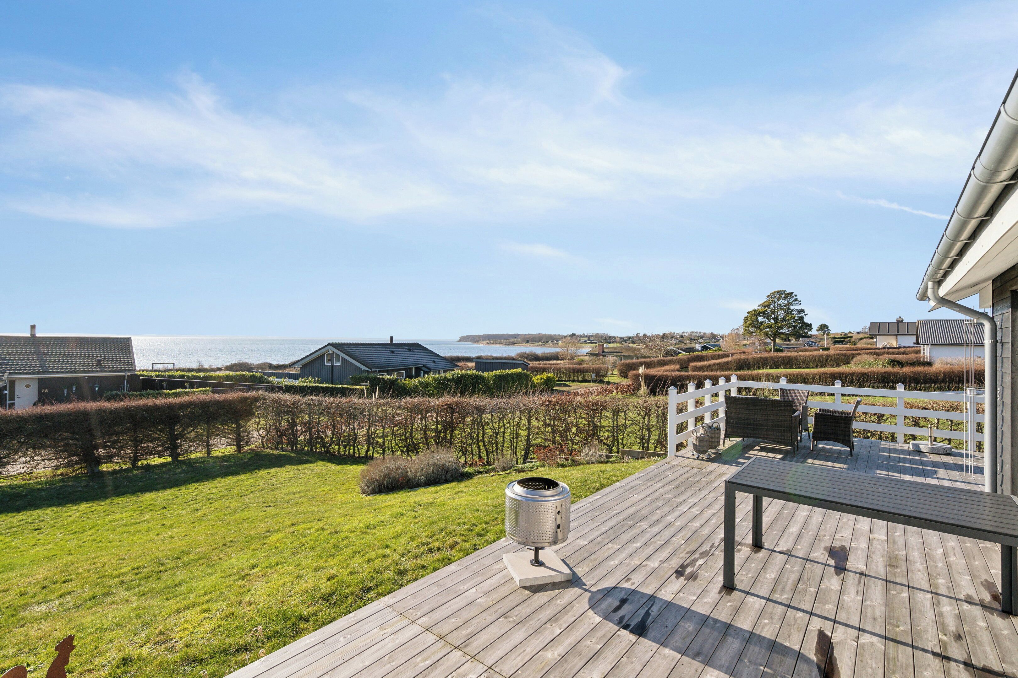 Une terrasse en bois avec un banc et un barbecue, donnant sur un jardin aménagé et un plan d’eau.