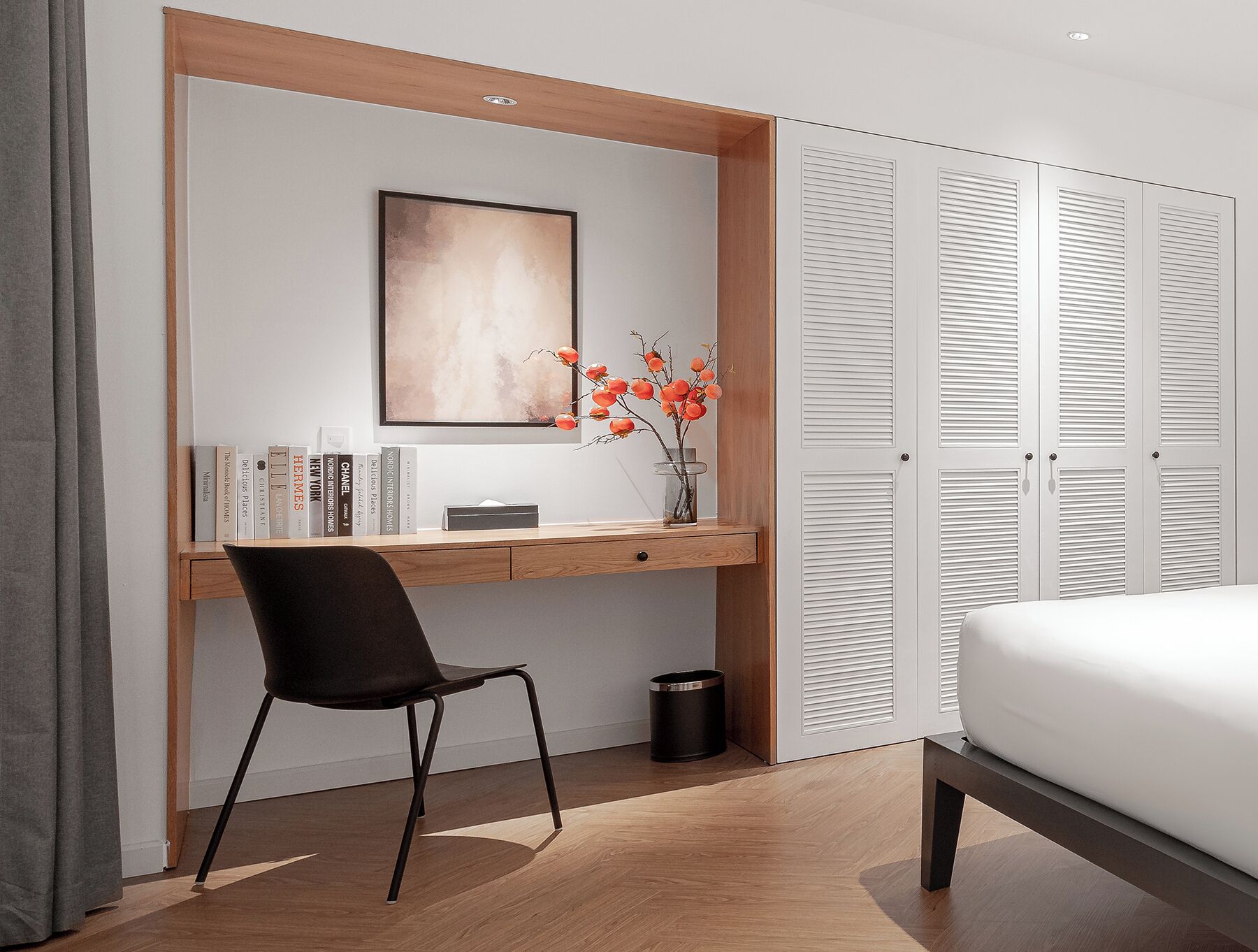 A modern bedroom with a wooden desk, a black chair, a bed, and a wardrobe with louvered doors.