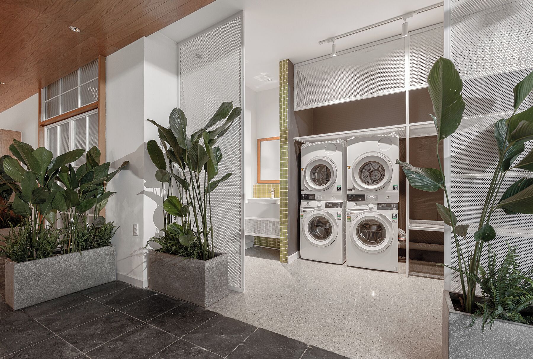 A laundry room with stacked washers and dryers, large potted plants, and a tiled floor.