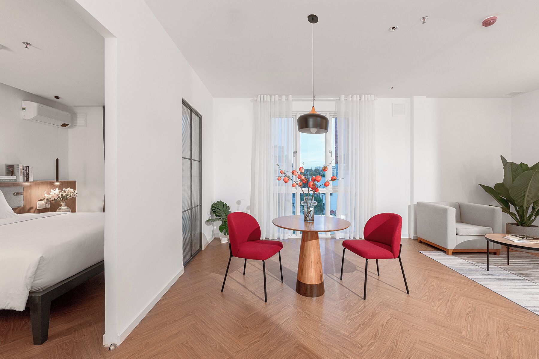 Een moderne woonkamer met een ronde houten tafel, twee rode stoelen, een grijze bank en een groot raam met gordijnen.