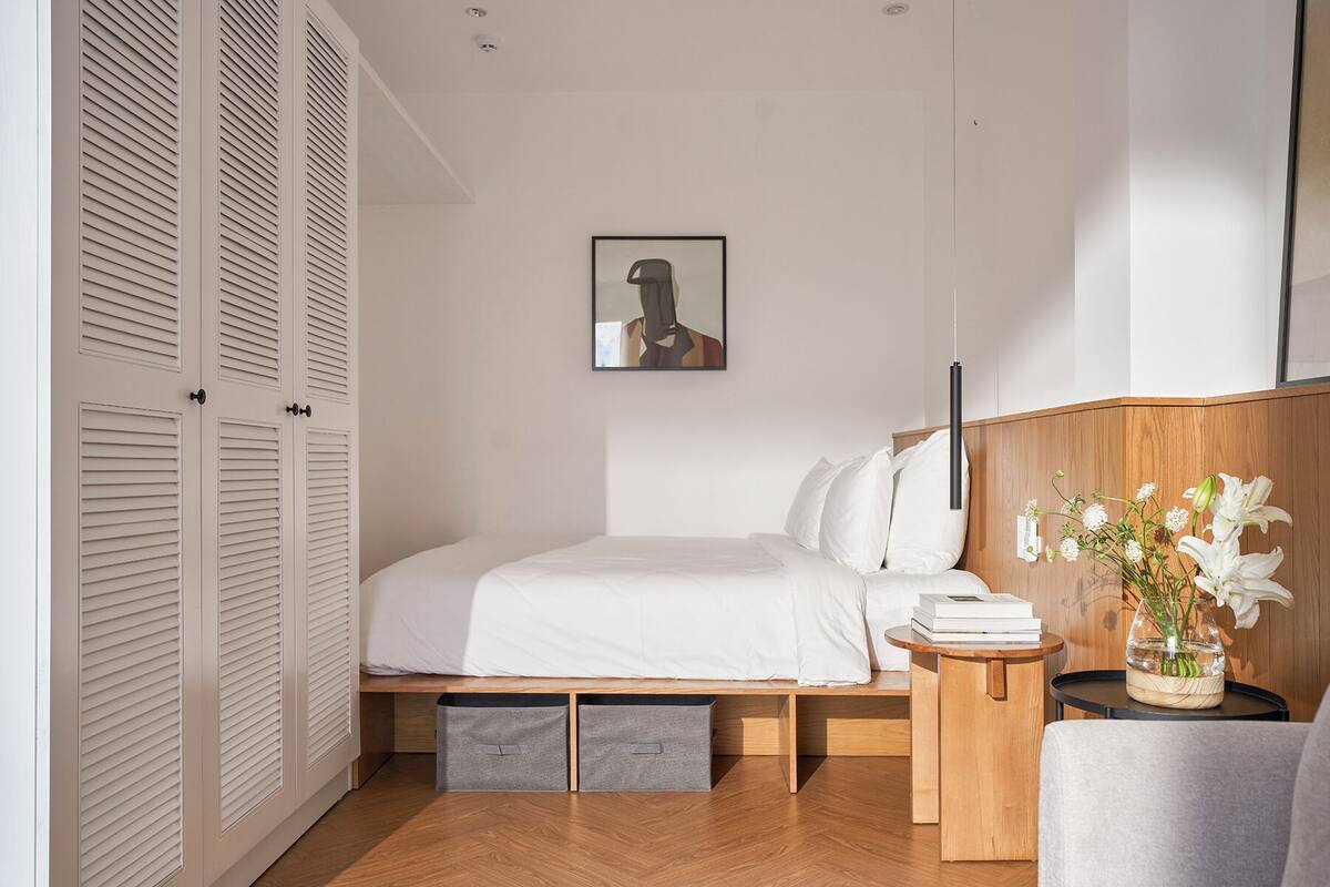 A bedroom with a bed, a bedside table with books, a vase with flowers, and a painting on the wall.