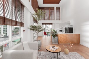 Un interior moderno con sala de estar, cocina y comedor. Hay ventanas grandes con persianas, un sofá, una mesa de centro y una encimera de cocina.