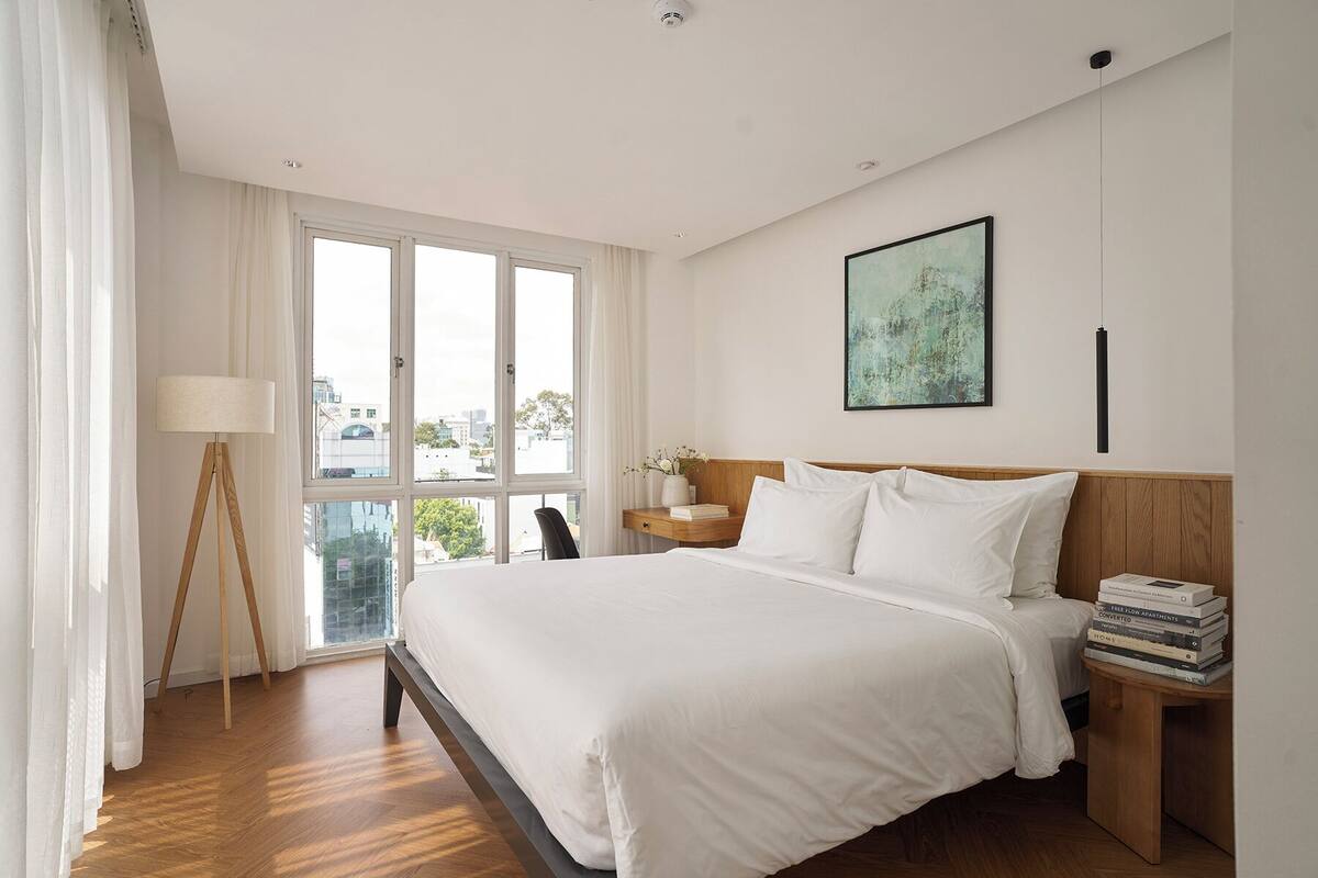 A hotel room with a large bed, a bedside table with books, a floor lamp, and a window with curtains.