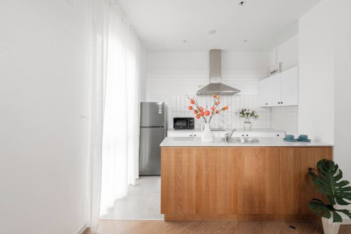 Una cocina moderna con muebles blancos, una isla de madera, electrodomésticos de acero inoxidable y un ventanal con cortinas translúcidas.