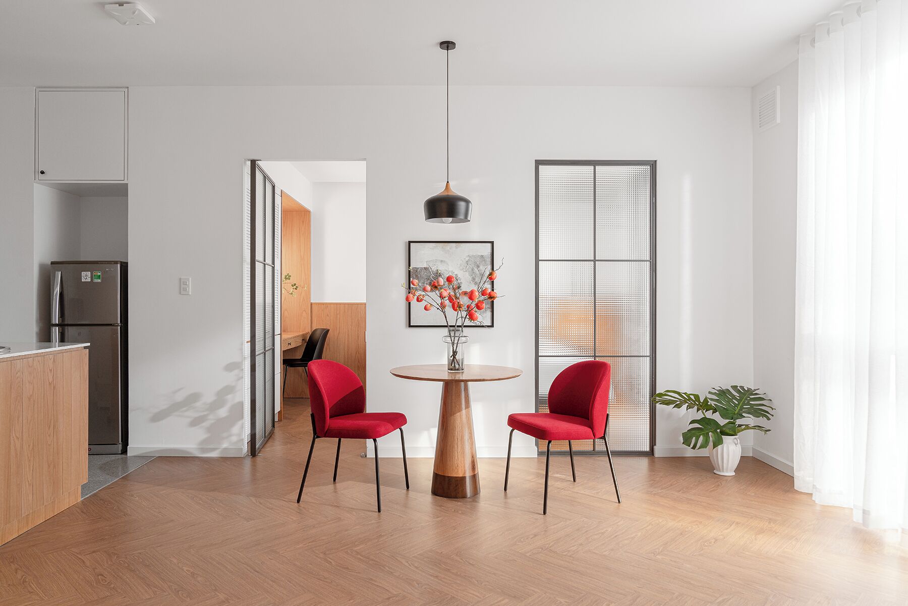 A modern kitchen with a refrigerator, wooden cabinets, and a dining area with two red chairs and a wooden table.