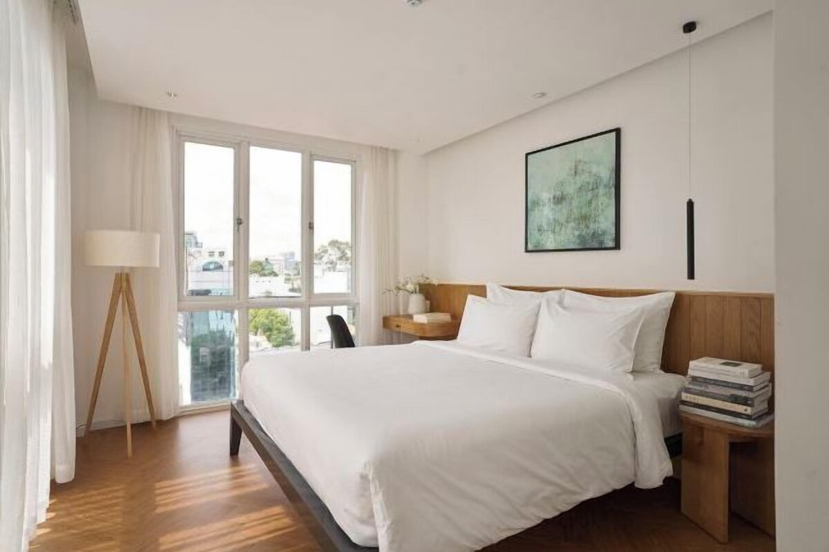 Habitación de hotel con una cama grande, una mesita de noche con libros, una ventana con cortinas y un cuadro en la pared.