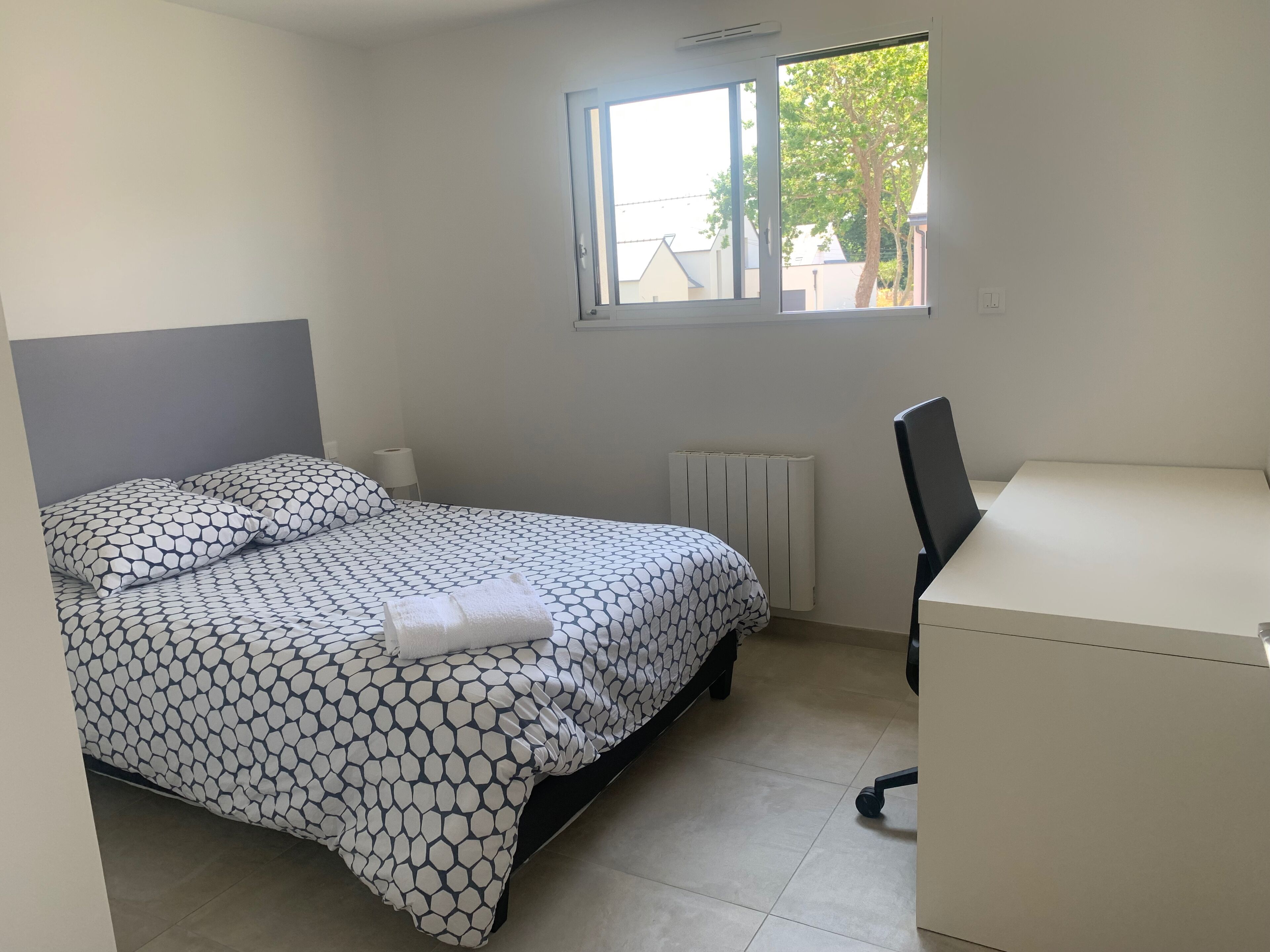 Un dormitorio con cama, escritorio, silla, radiador y ventana.
