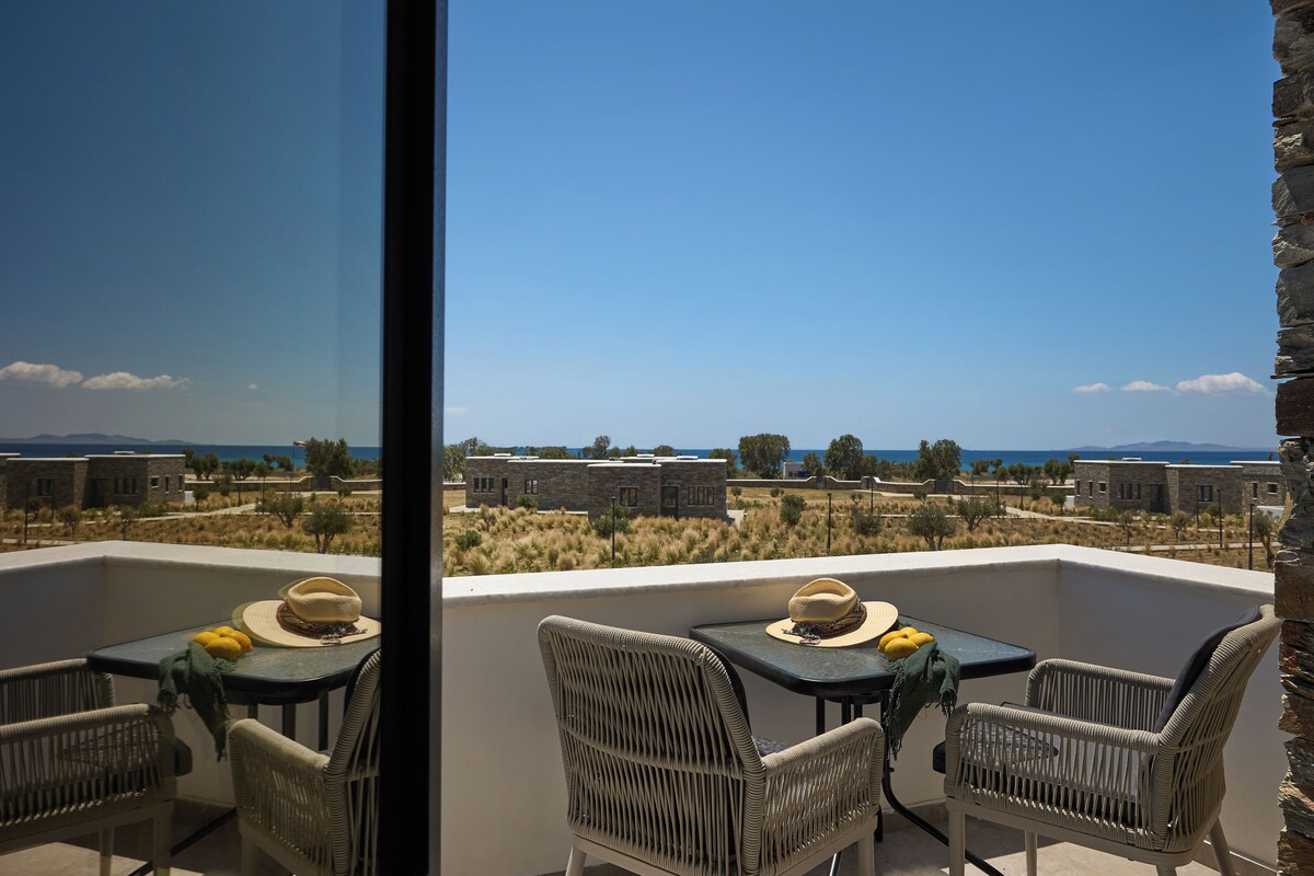 A balcony with wicker chairs and a table, overlooking a scenic landscape with buildings and a clear sky.