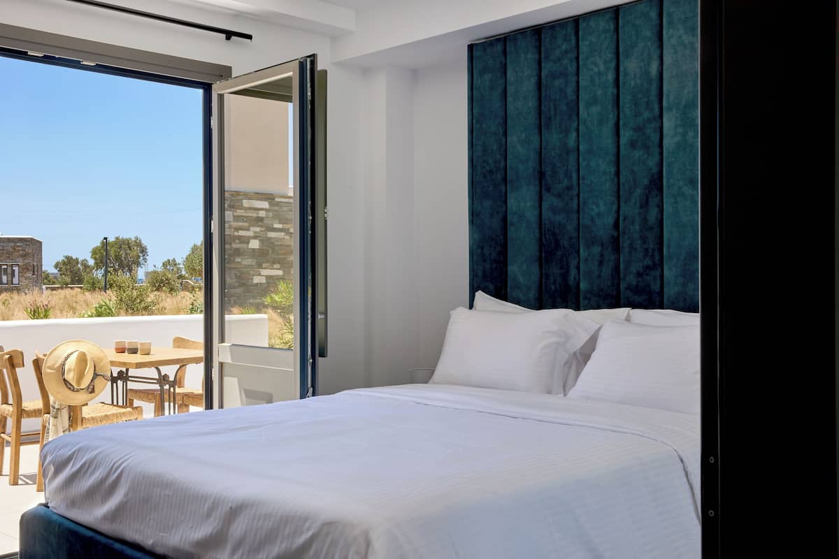 Habitación de hotel moderna con una cama grande, un balcón con mesa y sillas, y vistas al exterior.